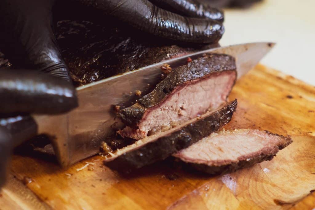 A juicy looking beef brisket is being sliced on a chopping block with a chef knife.