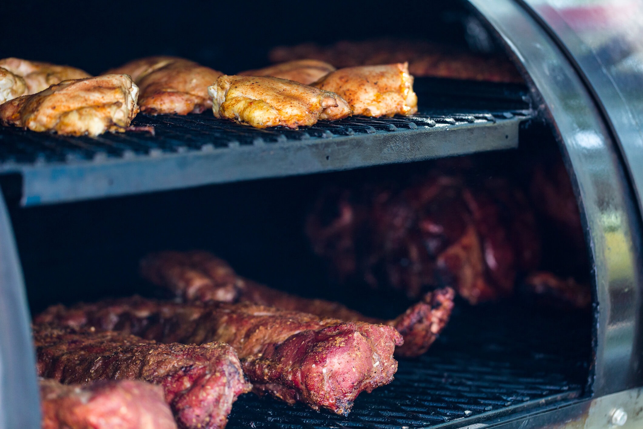 Smoker filled with seasoned chicken thighs on the top rack and pork ribs cooking on the bottom rack.