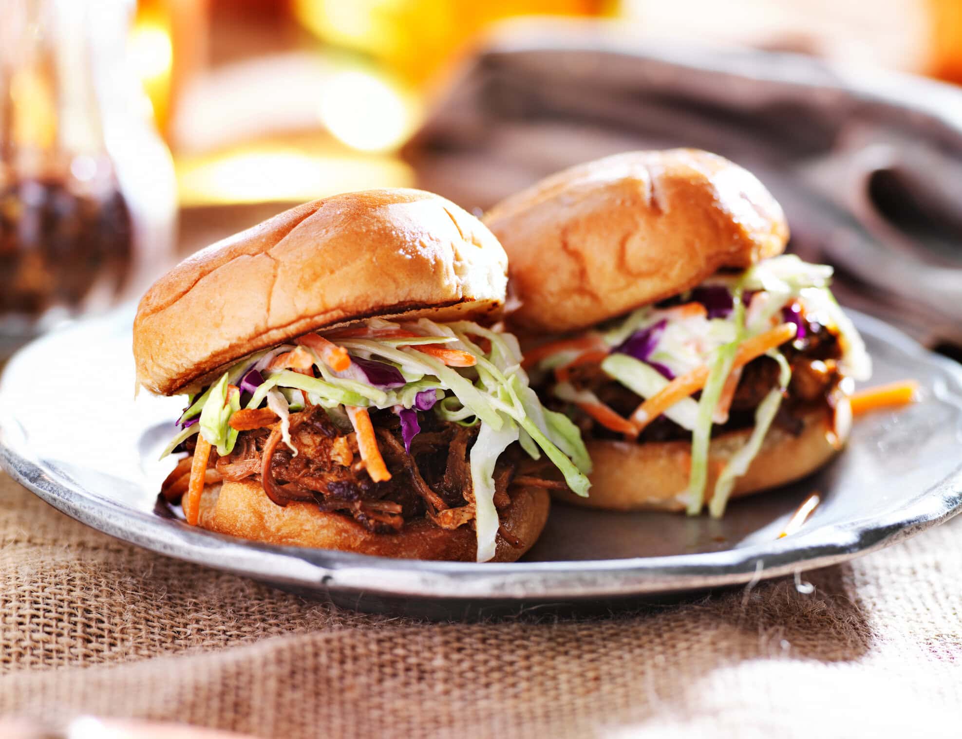 Two pulled pork sandwiches topped with fresh coleslaw on toasted buns, served on a metal plate with a rustic burlap table setting.