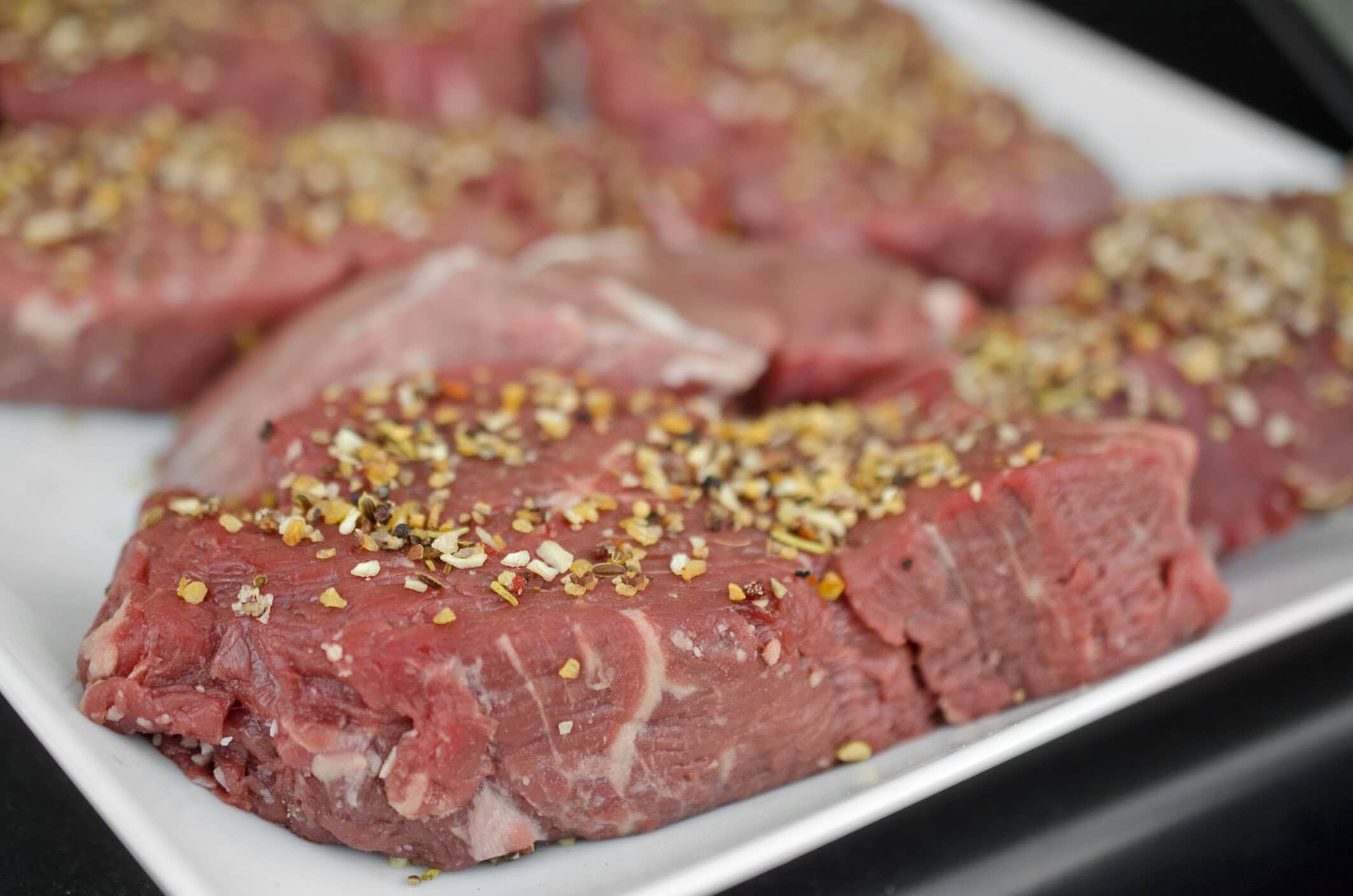 Raw pork steaks seasoned with cracked pepper and spices on a white plate.