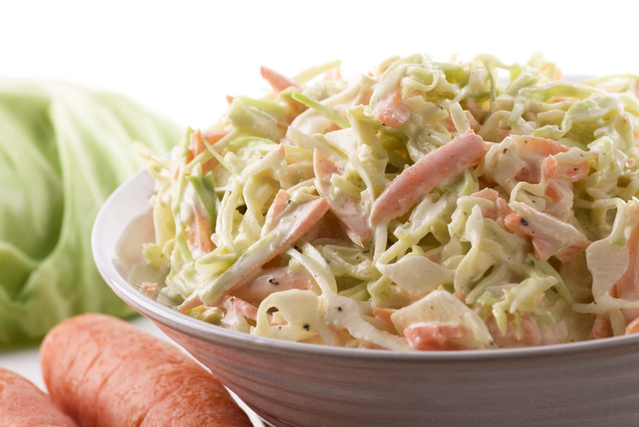 Close-up of creamy coleslaw with shredded cabbage and carrots in a bowl, served next to fresh carrots and cabbage.