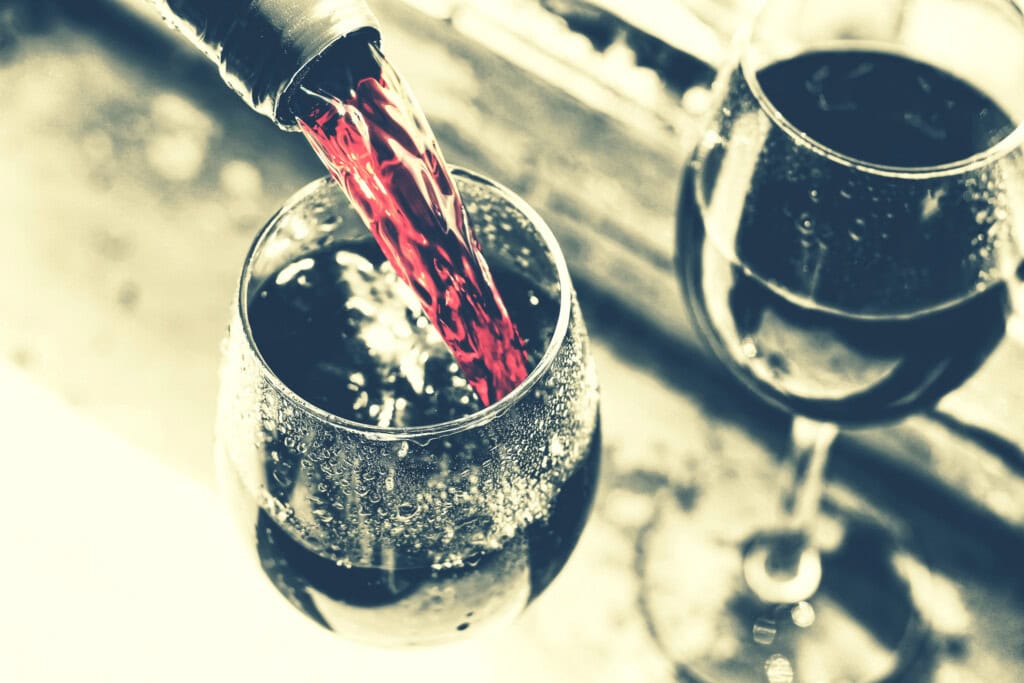 Red wine being poured into a glass with condensation, with a second filled wine glass in the background.