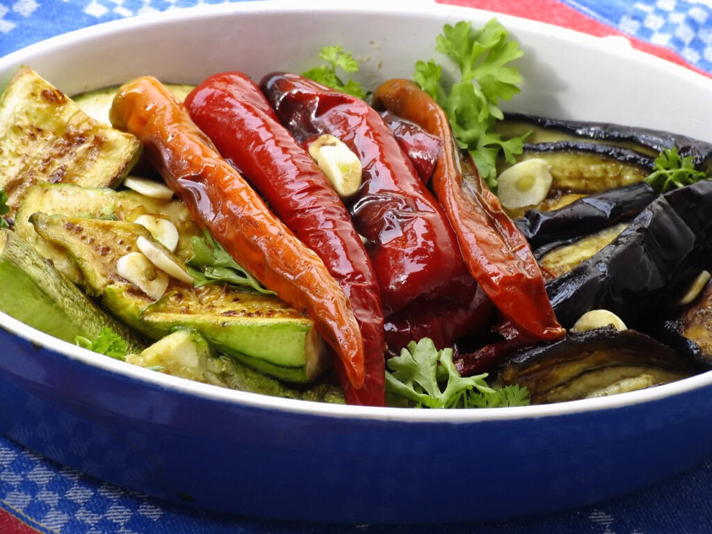 Grilled vegetables including zucchini, red peppers, and eggplant garnished with garlic and parsley in a serving dish.