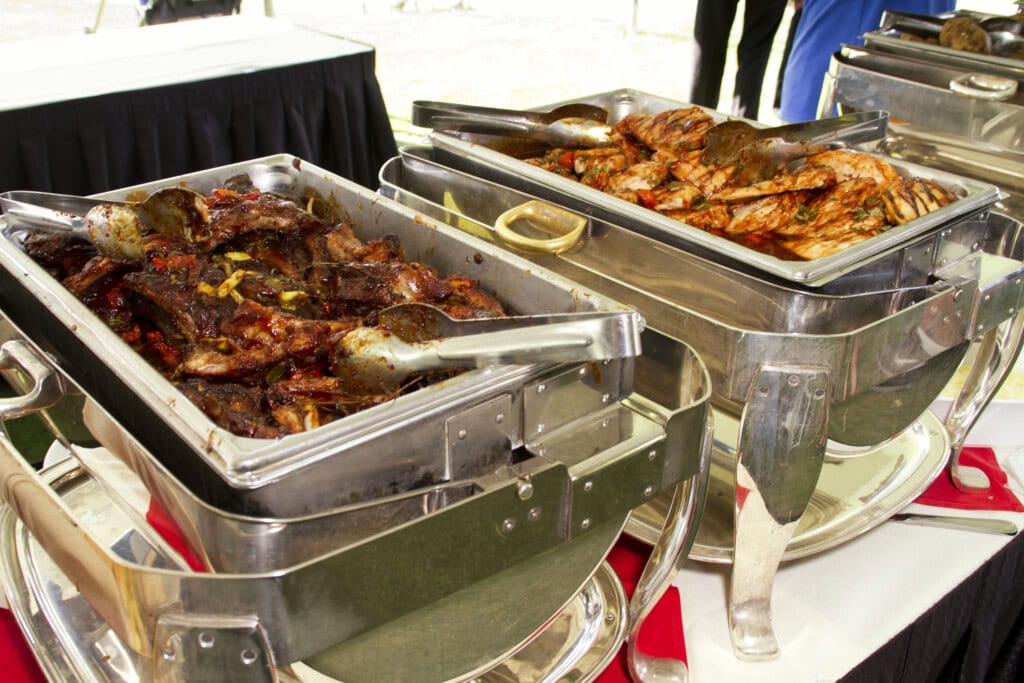 Buffet trays filled with barbecued meats, including ribs and grilled chicken, served with tongs at a catered event.