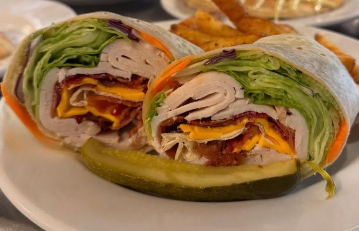 Smoked turkey wrap for lunch with bacon, cheddar cheese, mayo, tomato, and lettuce cut in half with a pickle spear on the side.